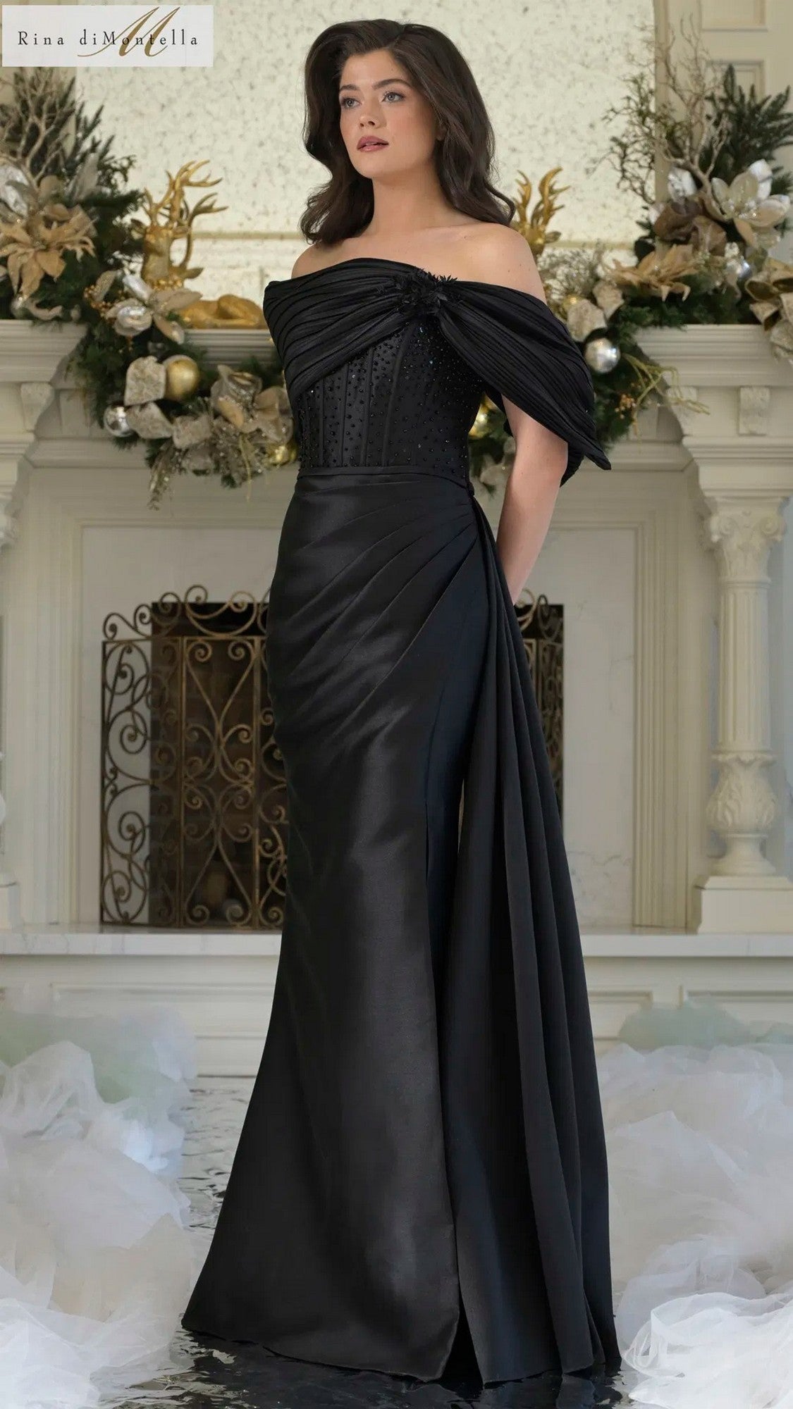 A woman stands indoors in the Long Formal Dress: Rina Di Montella RD3309, a black off-shoulder, floor-length gown with a fitted bodice and sheath skirt. Behind her is a white fireplace adorned with gold accents.